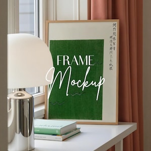 以下が含まれることがあります： 緑色の背景と鳥の群れがある額入りのアートプリント。 フレームは明るい木材です。 「FRAME Mockup」というテキストは白いスクリプトで書かれています。 白いランプと本が白いテーブルの上にあります。
