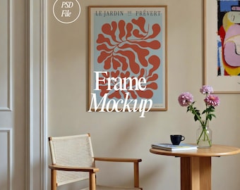 May include: A framed poster with a blue background and a red abstract design. The poster is titled "Le Jardin de Prévert". The poster is hanging on a light beige wall with a wooden frame. There is a wooden chair and a small round table with a book and a cup on it in the foreground.