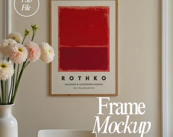 May include: A framed abstract painting with two red rectangles, one on top of the other, with the text "ROTHKO" and "SOLOMON R. GUGGENHEIM MUSEUM" below the painting. The frame is wooden and the painting is hanging on a beige wall. A white vase with flowers is on a wooden table with a book and a white chair.