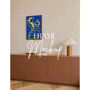 Op de afbeelding: Een ingelijst abstract bloemenkunstwerk met witte en groene accenten op een blauwe achtergrond hangt boven een bruine kast. Een houten lamp met een geplooide lampenkap staat op de kast. De woorden "FRAME" en "Mockup" zijn in witte letters geschreven.