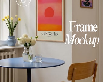 May include: A framed print of a sunset painting by Andy Warhol, with the title "Sunset" below the image. The print is hanging on a white wall in a room with a blue table and a wooden chair. There is a smaller framed print of a floral arrangement on the wall to the right of the larger print.