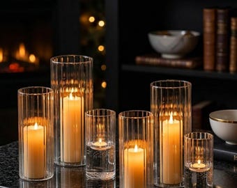 May include: A collection of clear glass candle holders of varying heights, each with a lit candle inside. The holders have vertical ribbed detailing and are arranged on a dark surface. The warm glow of the candles creates a cozy ambiance.