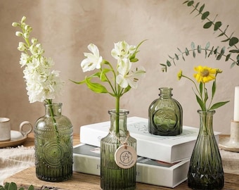 May include: Four decorative green glass vases of varying shapes and textures, each holding fresh flowers. The vases are arranged on a wooden surface with books in the background. One vase has a tag that says "ARTISAN LONDON".