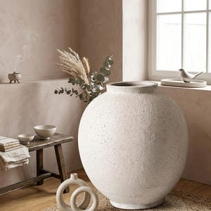 Könnte beinhalten: Eine große, kugelförmige, cremeweiße Keramikvase mit einer texturierten, gesprenkelten Oberfläche und einer weiten Öffnung. Die Vase steht auf einem gewebten Teppich, begleitet von einer kleineren runden Keramikskulptur, einer Holzbank und Zweigen.