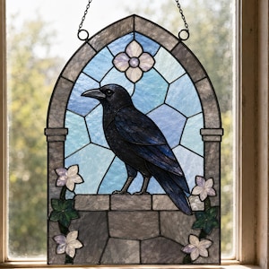 May include: A stained glass window hanging featuring a black raven perched on a stone ledge. The background consists of light blue geometric glass panels with a white flower at the top and green leaves with white flowers at the bottom corners.