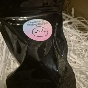 May include: A black, opaque, resealable pouch featuring a circular sticker with a gradient background ranging from blue to pink. The sticker displays the text "Mystery Dumplings" above a simple, black-lined illustration of a smiling dumpling character.