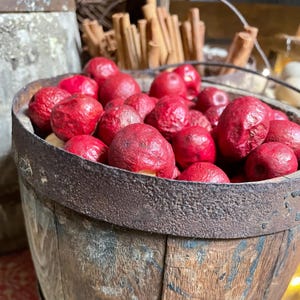 May include: A rustic wooden barrel filled with vibrant red decorative apples. The barrel has metal bands and a weathered appearance, suggesting a vintage aesthetic. Cinnamon sticks are visible in the background, adding to the autumnal theme.