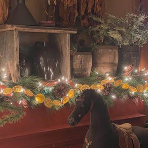 Puede incluir: Un caballo balanc&iacute;n marr&oacute;n oscuro con una silla de montar y brida color beige se encuentra frente a una chimenea de ladrillo rojo. La repisa est&aacute; decorada con una guirnalda de luces, pi&ntilde;as y rodajas de naranja secas. Detr&aacute;s del caballo hay barriles de madera r&uacute;sticos.