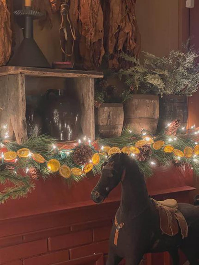 Puede incluir: Un caballo balanc&iacute;n marr&oacute;n oscuro con una silla de montar y brida color beige se encuentra frente a una chimenea de ladrillo rojo. La repisa est&aacute; decorada con una guirnalda de luces, pi&ntilde;as y rodajas de naranja secas. Detr&aacute;s del caballo hay barriles de madera r&uacute;sticos.
