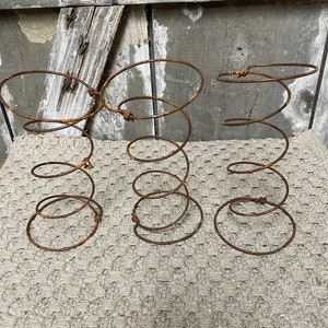 May include: Three rusty metal bed spring bases, each with a circular top and a coiled spring design. The bases are arranged in a row on a light brown knitted surface.
