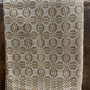 May include: A woven textile blanket featuring a repeating geometric pattern in mustard yellow and off-white. The fabric displays a textured, interlaced design with diamond and floral-like shapes distributed evenly across the entire surface area.