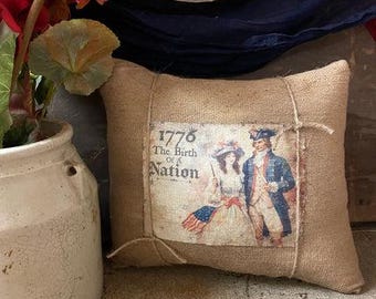 Puede incluir: Un cojín rectangular de arpillera con una impresión vintage que representa a una pareja con vestimenta colonial, el texto "1776 The Birth of a Nation" y una bandera estadounidense. El cojín está atado con hilo.