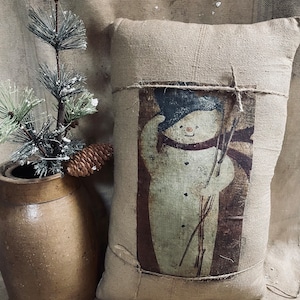 May include: A burlap pillow featuring a vintage-style snowman illustration. The snowman wears a black hat and a red scarf, holding sticks. The pillow is tied with twine and has a rustic, farmhouse aesthetic. A brown ceramic vase with a pine branch and pinecone is on the left.