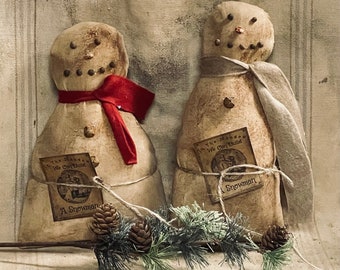 May include: Two primitive-style fabric snowman figures with distressed beige surfaces, dark pin-like eyes and mouths, and orange noses. One wears a red scarf, the other a grey scarf. Each has a tag reading In The Meadow We Can Build A Snowman.