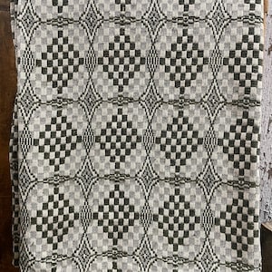 May include: A woven textile blanket featuring a repeating geometric pattern of dark olive green and off-white diamonds and checkered squares. The fabric displays a textured, interlaced weave structure with a consistent, symmetrical grid design throughout.