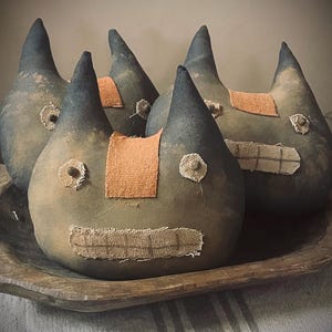May include: Four soft, stuffed fabric creatures with pointed ears, dark grey tops, and tan bodies. Each features a rectangular orange fabric patch on the forehead, two circular tan fabric eyes, and a wide, rectangular tan fabric mouth.