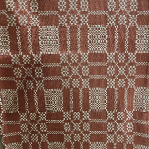 May include: A woven textile fabric featuring a repeating geometric pattern in terracotta orange and off-white. The design consists of alternating blocks of vertical and horizontal lines, creating a structured, grid-like appearance across the surface.