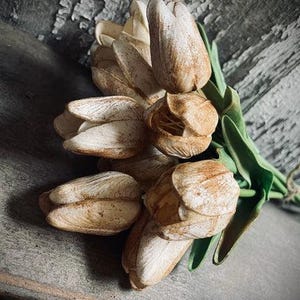 May include: A bouquet of faux tulips with cream-colored petals and brown accents. The artificial flowers have green leaves and are tied together with twine. The background is a weathered, gray wooden surface.