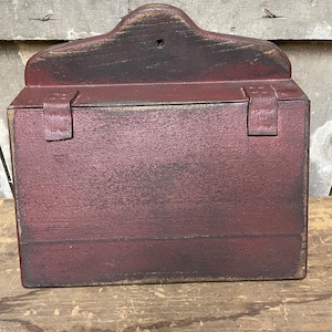 May include: A rectangular, burgundy-colored wooden box with a decorative top and two leather-like straps. The box has a distressed finish, suggesting an antique or rustic style. The box is sitting on a wooden surface.