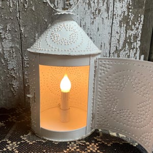 May include: A white metal lantern with a decorative punched star pattern and a hinged door. The lantern has a handle and a lit candle-shaped light inside, casting a warm glow. The background is a weathered wooden surface.