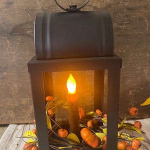 May include: Black metal lantern with a handle, featuring a lit, faux candle. The lantern is decorated with a garland of orange pumpkins and yellow leaves, creating a fall or Halloween theme. The background is a rustic wooden surface.