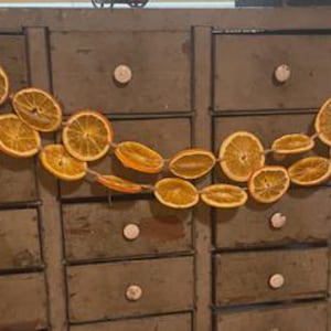 Puede incluir: Una guirnalda decorativa de rodajas de naranja secas ensartadas en hilo. Las rodajas de naranja son de un color dorado c&aacute;lido y est&aacute;n dispuestas en un patr&oacute;n fluido. La guirnalda se exhibe contra un mueble antiguo con m&uacute;ltiples cajones.