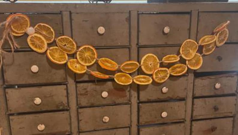Puede incluir: Una guirnalda decorativa de rodajas de naranja secas ensartadas en hilo. Las rodajas de naranja son de un color dorado c&aacute;lido y est&aacute;n dispuestas en un patr&oacute;n fluido. La guirnalda se exhibe contra un mueble antiguo con m&uacute;ltiples cajones.
