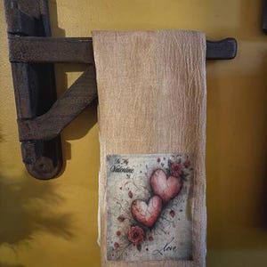 May include: A beige Valentine's Day towel with a decorative print of two red hearts and roses. The towel is hanging on a dark brown wooden towel rack. The print includes the words "Be My Valentine" and "Love".