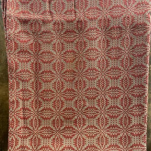 May include: A woven textile blanket featuring a repeating geometric pattern in terracotta orange and natural beige. The fabric displays a textured, interlaced weave structure with a consistent diamond-like motif across the entire surface area.