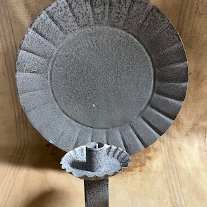 May include: A gray metal candle holder with a large, round backplate and a smaller, scalloped candle cup. The backplate has a textured surface and a decorative, fluted edge. The candle cup is attached to a rectangular base.