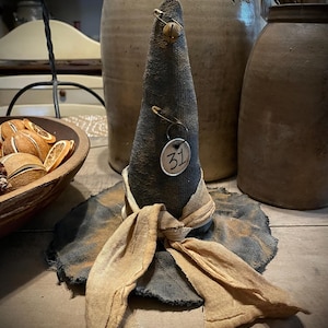 May include: A handcrafted Halloween witch hat with a cone shape, made of dark gray fabric with a tan brim and scarf. A small bell and a tag with the number 31 are attached. The hat is displayed on a wooden surface.