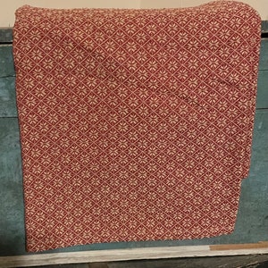 May include: A folded red and cream-colored patterned textile. The fabric features a repeating diamond pattern with floral-like designs. The textile is draped over a dark blue surface, with a portion of the fabric hanging over the edge.