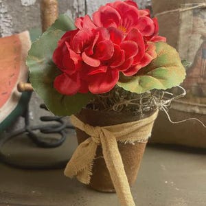May include: A small, rustic flower pot filled with vibrant red geraniums and green leaves. The pot is wrapped with a tan burlap ribbon. The arrangement evokes a farmhouse or cottagecore aesthetic.