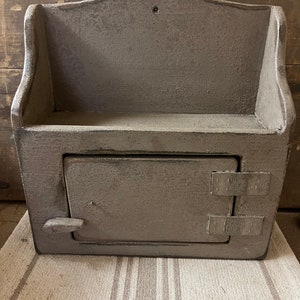 May include: A gray wooden wall-mounted cabinet with a decorative top and a small door. The cabinet has a distressed finish, giving it a rustic appearance. The door is secured with two rectangular latches and a small handle.