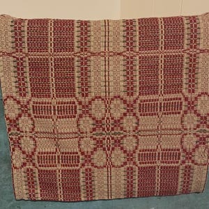 May include: A square textile piece featuring a woven geometric pattern in deep red and natural beige tones. The design consists of repeating blocks and grid-like structures with subtle dark green accents integrated into the woven fabric texture.