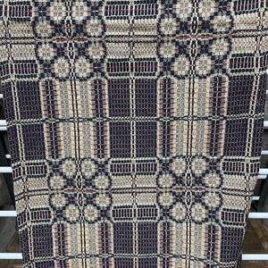 May include: A woven textile with a geometric pattern in navy blue, beige, and red. The design features repeating squares and floral-like shapes. The fabric appears to be hanging, showcasing its texture and pattern.