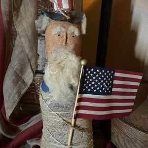 May include: A handmade patriotic doll dressed in red, white, and blue with a burlap-wrapped body and a white beard. The doll is holding a small American flag.