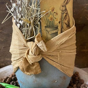 May include: A gray stone egg wrapped in a brown fabric with a sprig of white blossoms and a vintage card featuring a rabbit and daffodils with the text "Spring Thyme".