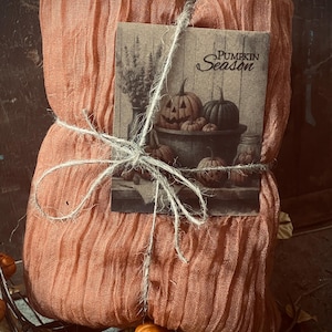 May include: A bundle of peach-colored fabric tied with twine, featuring a card with the text "Pumpkin Season" and a sepia-toned illustration of pumpkins. The fabric has a crinkled texture, suggesting a decorative item.