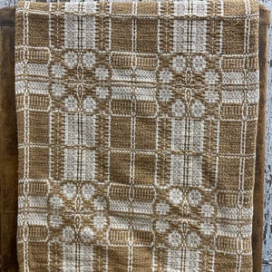 May include: A rectangular woven textile featuring a repeating geometric pattern in shades of golden brown and off-white. The fabric displays a complex grid-like structure with alternating blocks of solid color and intricate, textured weave details.