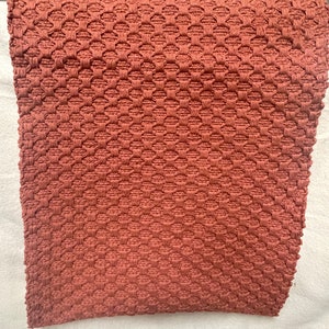 May include: A rust-colored woven rug with a textured diamond pattern. The rug is made of a soft, natural material and has a subtle sheen.