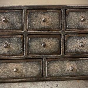 May include: A small, rustic wooden cabinet with nine drawers. The cabinet has a distressed, weathered finish in shades of gray and brown. Each drawer has a small, round metal knob. The cabinet is designed for storage.