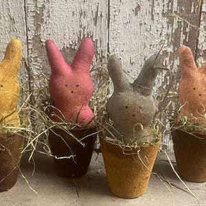 May include: Four Easter bunny decorations in terracotta pots. The bunnies are in shades of yellow, pink, gray, and orange. Each bunny is surrounded by dried grass. The pots are a rustic brown color.
