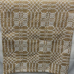 May include: A woven textile with a repeating geometric pattern in gold and white. The fabric appears to be folded over, revealing the intricate design. The texture suggests a soft, possibly natural fiber material.