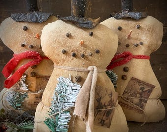 May include: Three snowman dolls with tan bodies, black top hats, and red scarves. Each snowman has peppercorn eyes and a carrot nose. They are decorated with small tags and sprigs of faux greenery, creating a rustic winter theme.