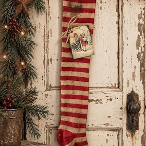 May include: A long Christmas stocking featuring horizontal red and beige stripes with a distressed, weathered texture. A rectangular paper tag depicting two children playing in the snow is tied to the stocking with a piece of brown twine.