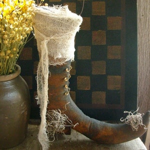 May include: A rustic, handcrafted witch boot decoration. The boot is brown with gold-colored accents and a pointed toe. It is wrapped in a cream-colored gauze and adorned with decorative moss. A wire loop is attached for hanging. A vase of dried yellow flowers is in the background.