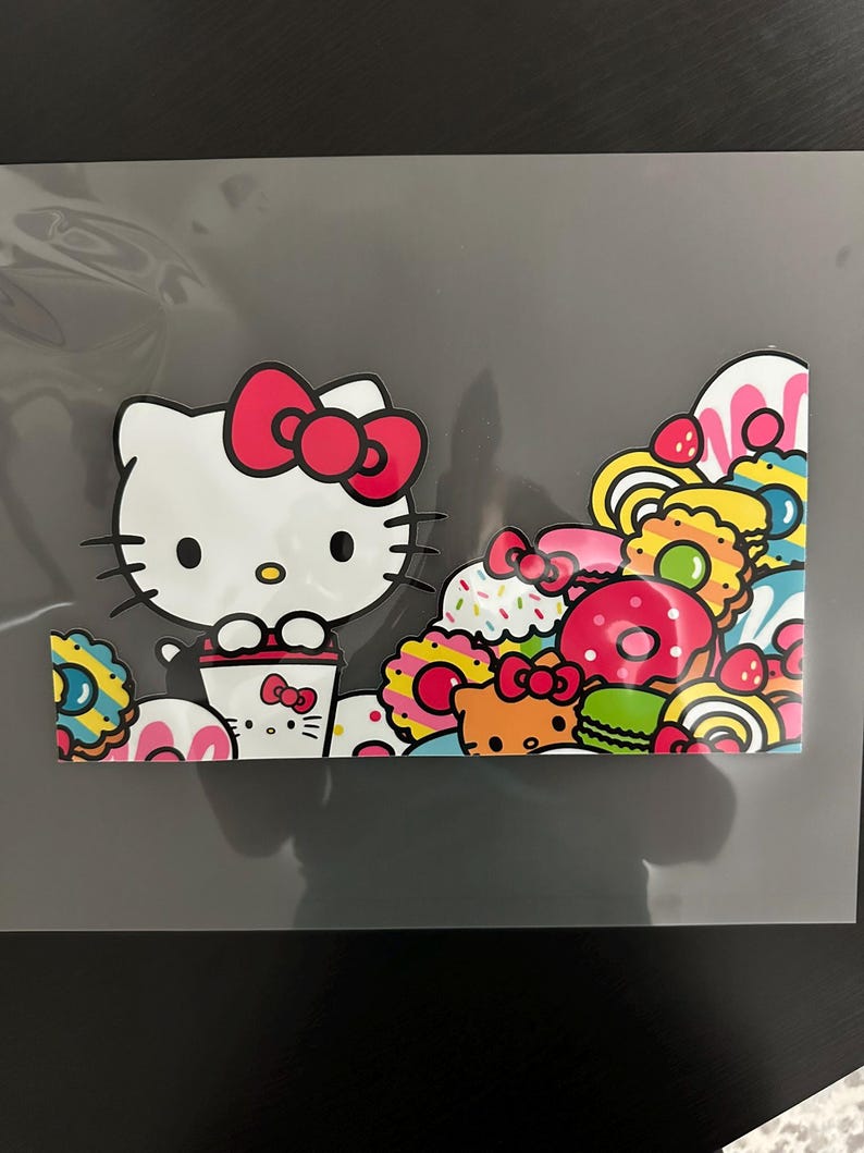 K&ouml;nnte beinhalten: Ein wei&szlig;er Hello Kitty Charakter mit einer roten Schleife h&auml;lt eine Tasse Kaffee. Der Hintergrund ist gef&uuml;llt mit bunten Donuts, Cupcakes und anderen s&uuml;&szlig;en Leckereien.