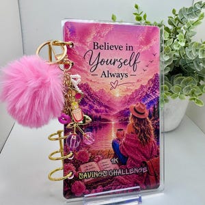 May include: A pink and gold spiral-bound savings challenge notebook with the text "Believe in Yourself - Always." The notebook features a scenic illustration and a pink pom-pom keychain. The notebook is designed to help track savings goals.