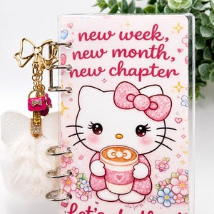 Könnte beinhalten: Ein rosa-weißer Planer mit Hello Kitty-Design, mit dem Text "new week, new month, new chapter" und "Let's do this!!". Enthält einen goldenen Schleifenanhänger, einen rosa Handtaschenanhänger und einen weißen flauschigen Anhänger.